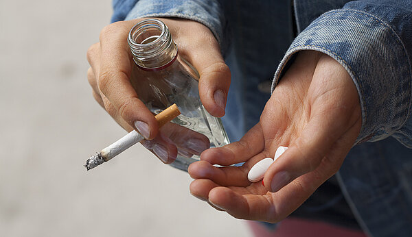 Zwei Hände halten eine Zigarette, eine kleine Flasche und diverse Tabletten