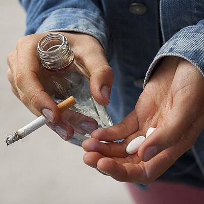 Zwei Hände halten eine Zigarette, eine kleine Flasche und diverse Tabletten