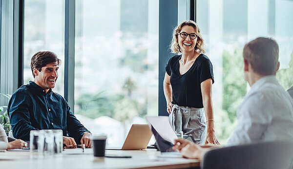 Drei Personen an einem Tisch in einem Raum, eine vierte steht und lacht