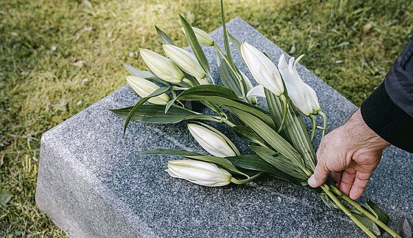 Eine Hand legt einen Lilienstrauß auf einen Grabstein