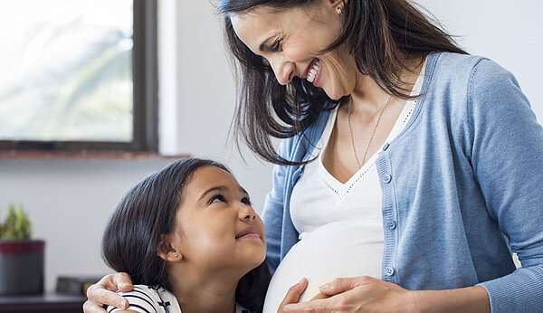 Kind hält schwangerer Frau die Hand auf den Bauch. Beide lächeln.