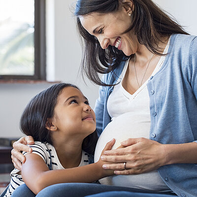 Kind hält schwangerer Frau die Hand auf den Bauch. Beide lächeln.