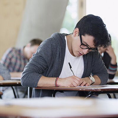 Mehrere junge Menschen sitzen in einem Raum an Einzeltischen und schreiben