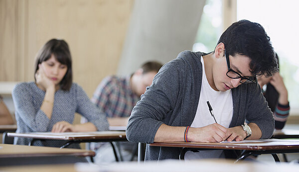 Mehrere junge Menschen sitzen in einem Raum an Einzeltischen und schreiben