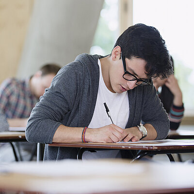 Mehrere junge Menschen sitzen in einem Raum an Einzeltischen und schreiben