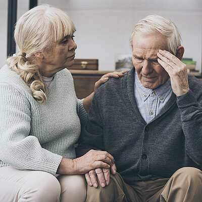 Eine weibliche Seniorin sitzt neben einem männlichen Senior und hat ihm die Hand auf die Schulter gelegt